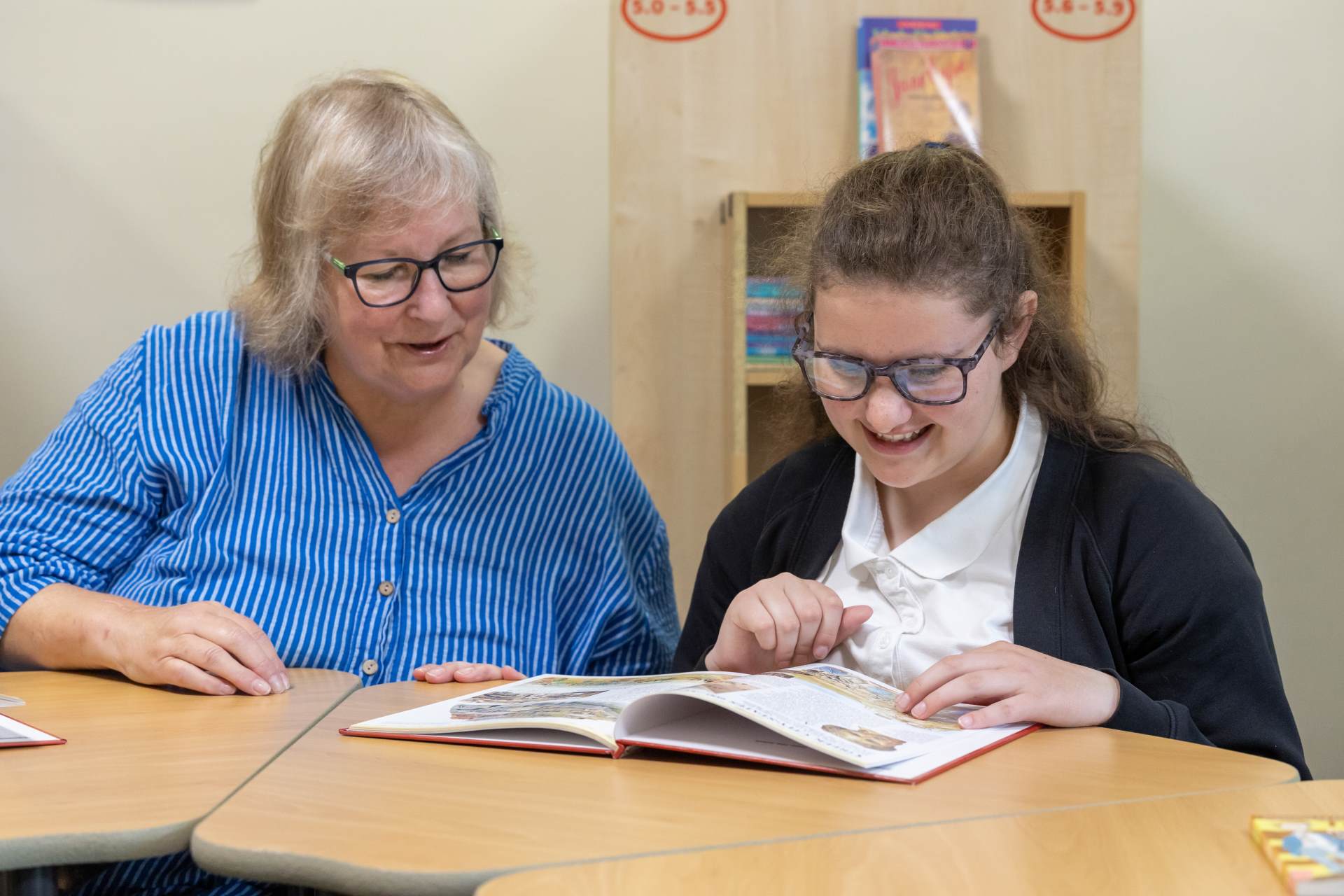 Trustee Alison with students at Fosseway School a woman with blonde/white hair and glasses and wearing a blue striped shirt is sitting next to a girl in a navy blue uniform. they are both looking at a book