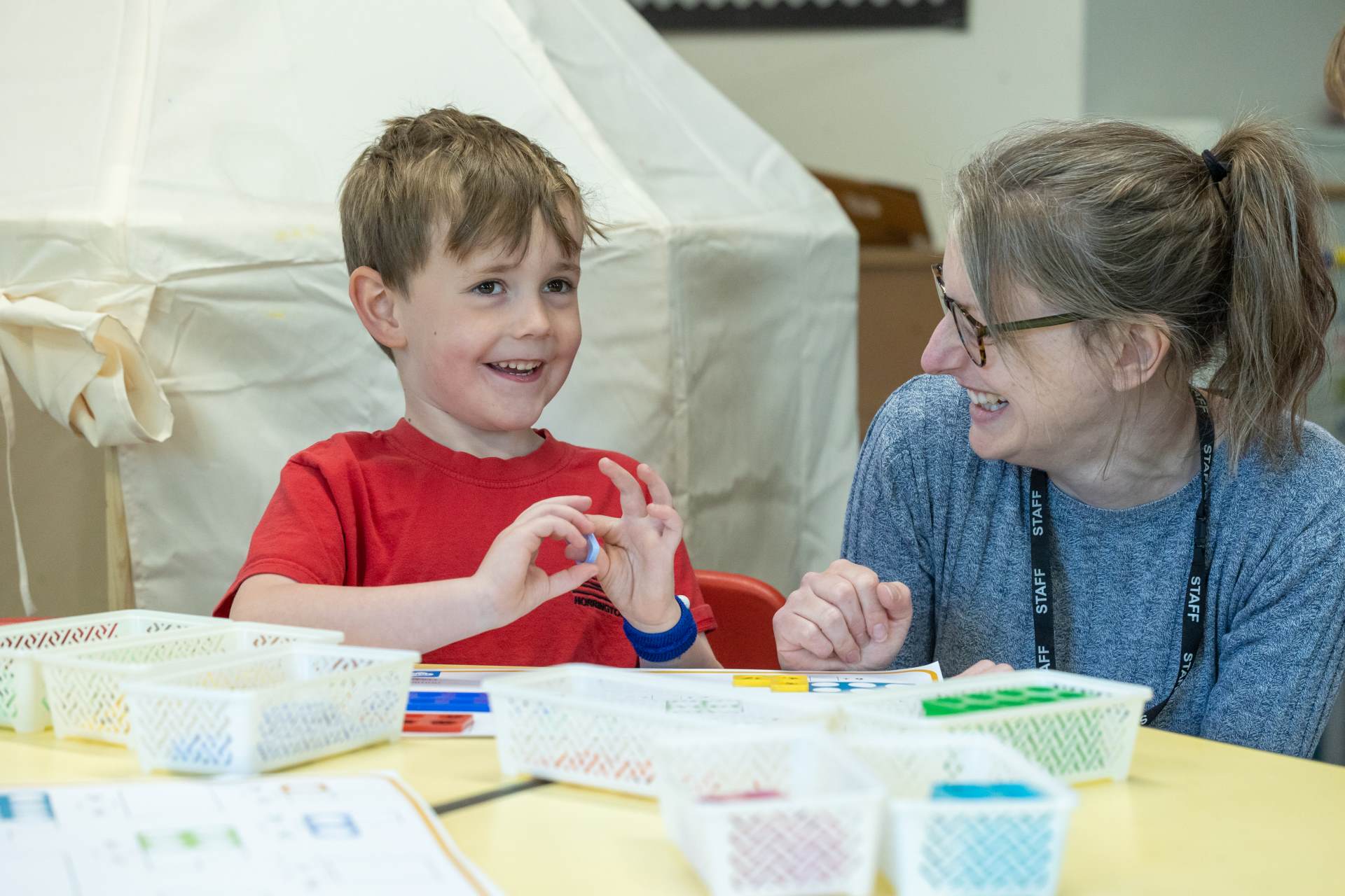 a young boy wearing a red tshirt is in a classroom with his female teacher. They have some plastic boxes in front of them