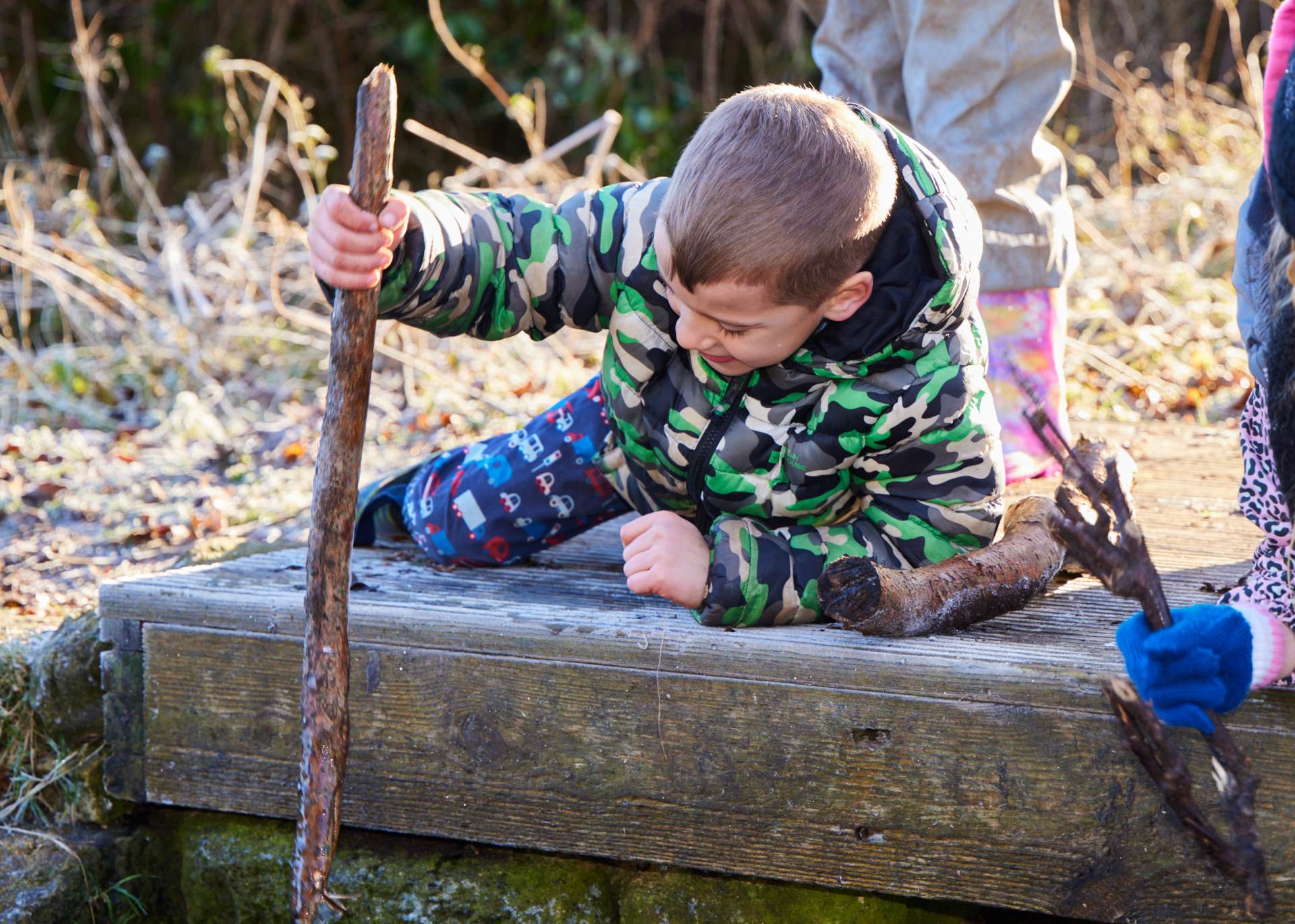 a young boy is wearing outdoor clothing and is pictured lying on his side. He has a wooden stick and is using that to poking a body of water