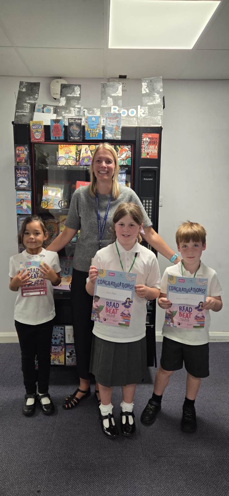 Kelly Sessions at Castle Primary School a woman with blonde hair and wearing a grey top is standing in front of a bookcase. she is smiling with three school pupils in front of her. They are all wearing white tops and grey skirts or trousers or leggings.